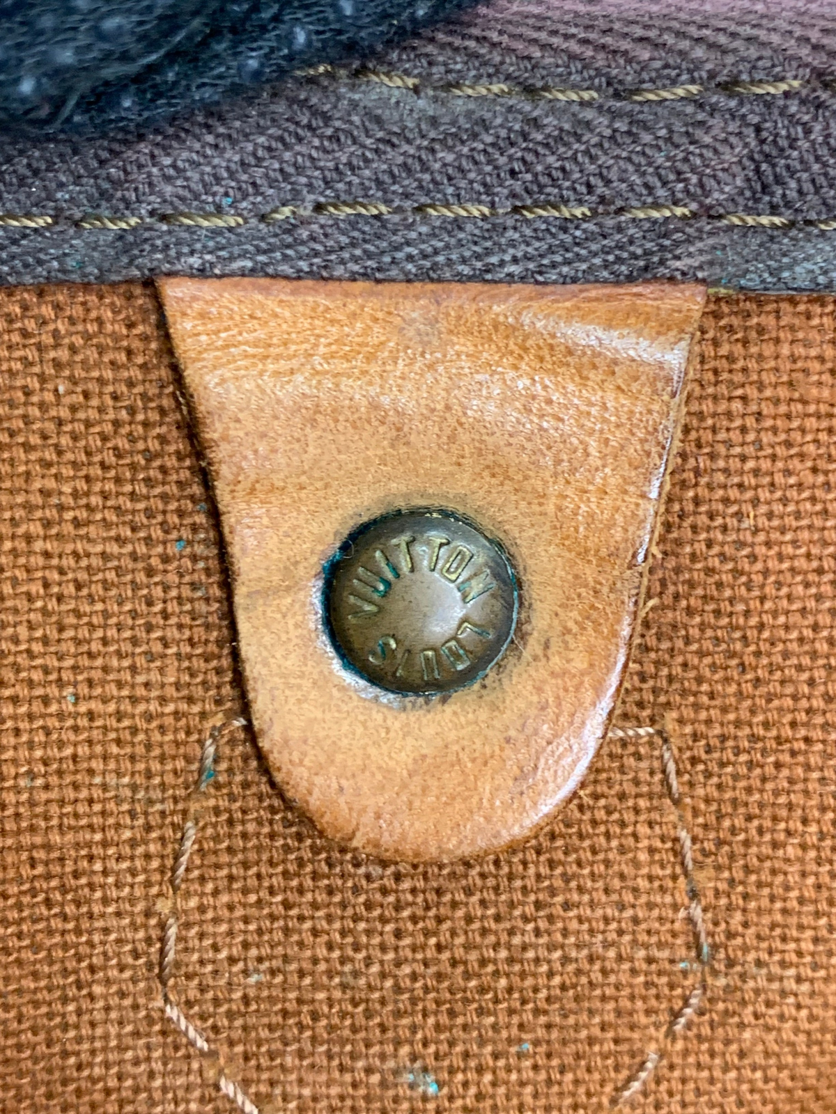 Close-up of a Louis Vuitton monogram canvas duffel bag featuring a branded brass rivet on a tan leather tab.
