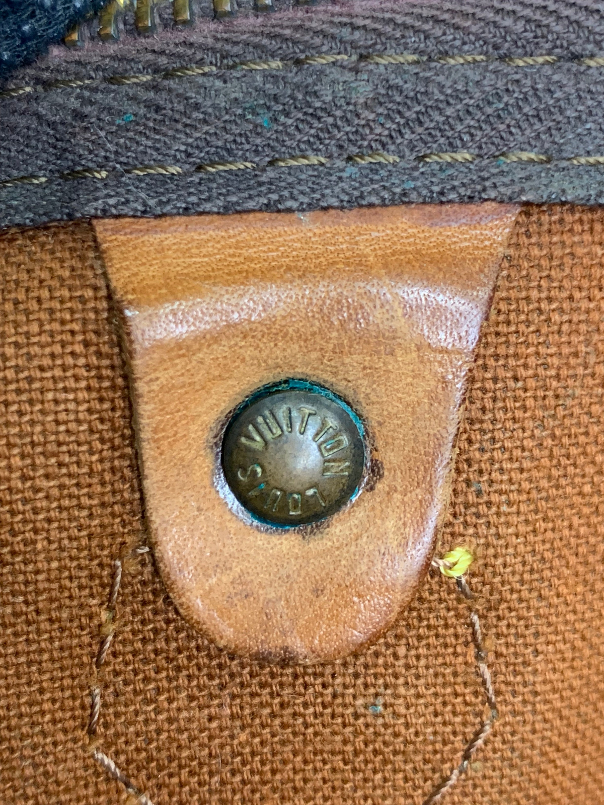 Close-up of a Louis Vuitton monogram duffel bag featuring a branded brass rivet on a tan leather tab.